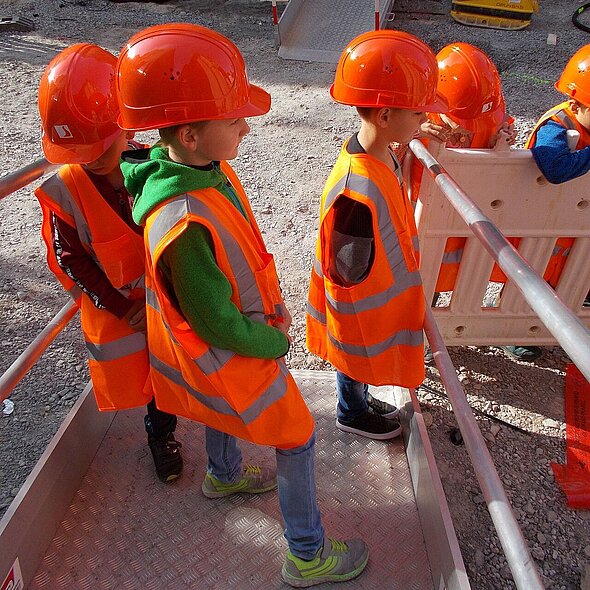 Vier Kinder mit Bauhelmen schauen einer Baustelle zu